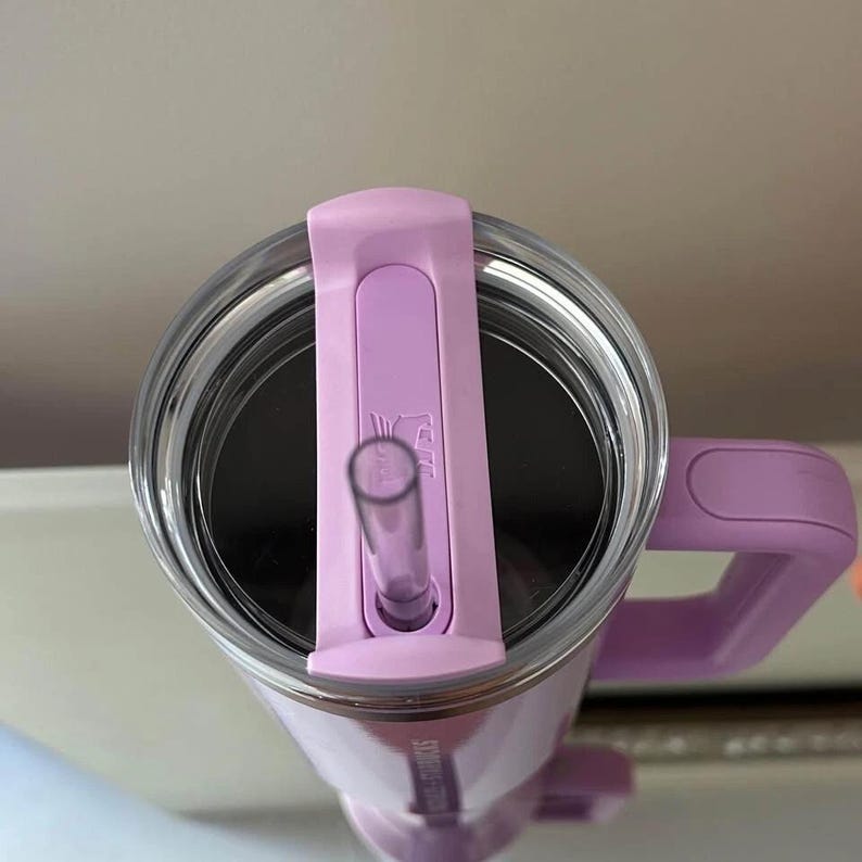 May include: A lavender-colored travel mug with a clear straw and a matching handle. The mug has a clear lid with a sliding closure for the straw. The mug has the words "STAINLESS STEEL" printed on the side.