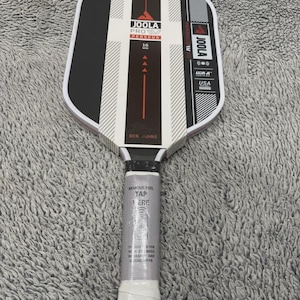 May include: A new Joola Pro Persues pickleball paddle with a black and white design. The paddle has a white grip and the text "REMOVE FOIL TAP HERE". The paddle is USA approved.