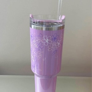 May include: A lavender tumbler with a clear lid and a clear straw. The tumbler features a floral design in white and purple. The tumbler is made of stainless steel and is approximately 9 inches tall.