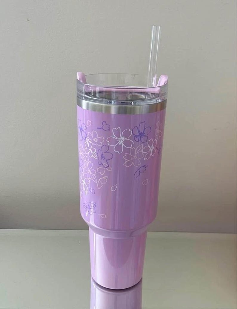 May include: A lavender tumbler with a clear lid and a clear straw. The tumbler features a floral design in white and purple. The tumbler is made of stainless steel and is approximately 9 inches tall.