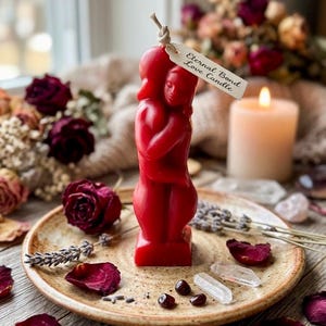 May include: A red candle shaped as two figures embracing, with a tag reading "Eternal Bond Love Candle." The candle is on a plate with rose petals and lavender. A lit candle and dried flowers are in the background.