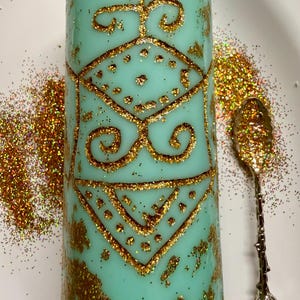 May include: A turquoise candle with gold glitter and a decorative gold pattern. An ornate spoon and scattered gold glitter sit beside the candle on a white surface. The candle features geometric shapes and swirls. The candle is cylindrical in shape.