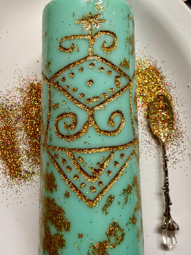 May include: A turquoise candle with gold glitter and a decorative gold pattern. An ornate spoon and scattered gold glitter sit beside the candle on a white surface. The candle features geometric shapes and swirls. The candle is cylindrical in shape.