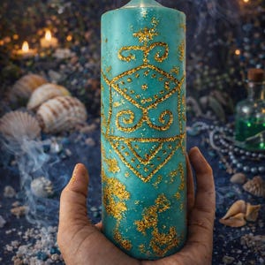 May include: A lit, turquoise pillar candle with gold glitter detailing. The candle features a geometric design and is held in a hand. The background includes seashells, crystals, and a blurred, dark blue backdrop.