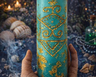 May include: A lit, turquoise pillar candle with gold glitter detailing. The candle features a geometric design and is held in a hand. The background includes seashells, crystals, and a blurred, dark blue backdrop.