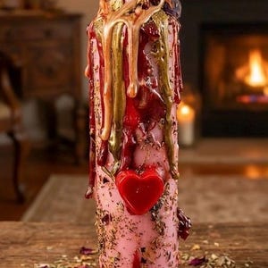 May include: A pink pillar candle with a red heart and gold and red wax drips. The candle is decorated with dried herbs and rose petals. The candle is on a wooden surface.