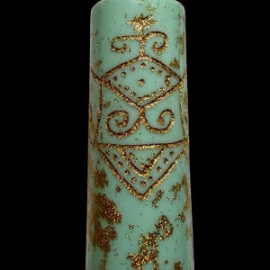 May include: A tall, mint-green pillar candle with gold glitter accents and a gold geometric design. The candle has a textured surface and a white wick. The design includes a star, dots, and swirls. The background is black.