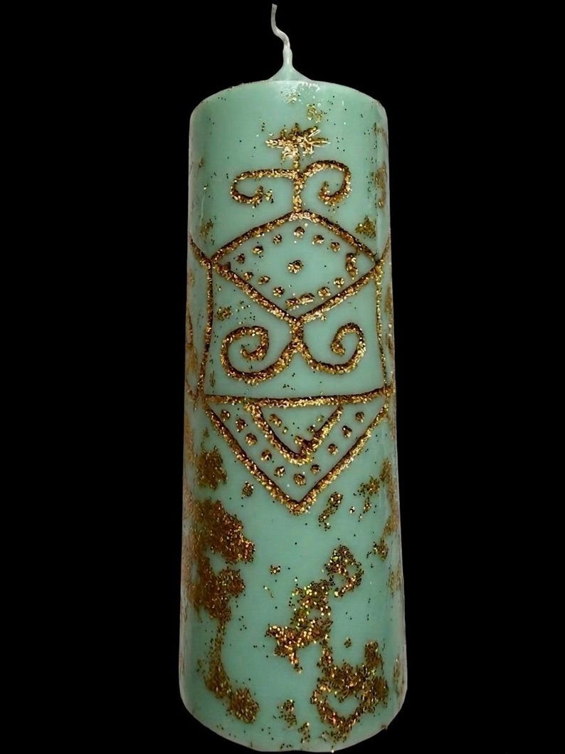 May include: A tall, mint-green pillar candle with gold glitter accents and a gold geometric design. The candle has a textured surface and a white wick. The design includes a star, dots, and swirls. The background is black.