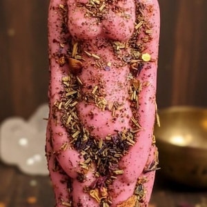 May include: A pink candle shaped like a female form, decorated with dried herbs and flowers. The candle has a wick and stands on a dark wooden surface. The overall aesthetic is rustic.