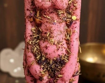 May include: A pink candle shaped like a female form, decorated with dried herbs and flowers. The candle has a wick and stands on a dark wooden surface. The overall aesthetic is rustic.