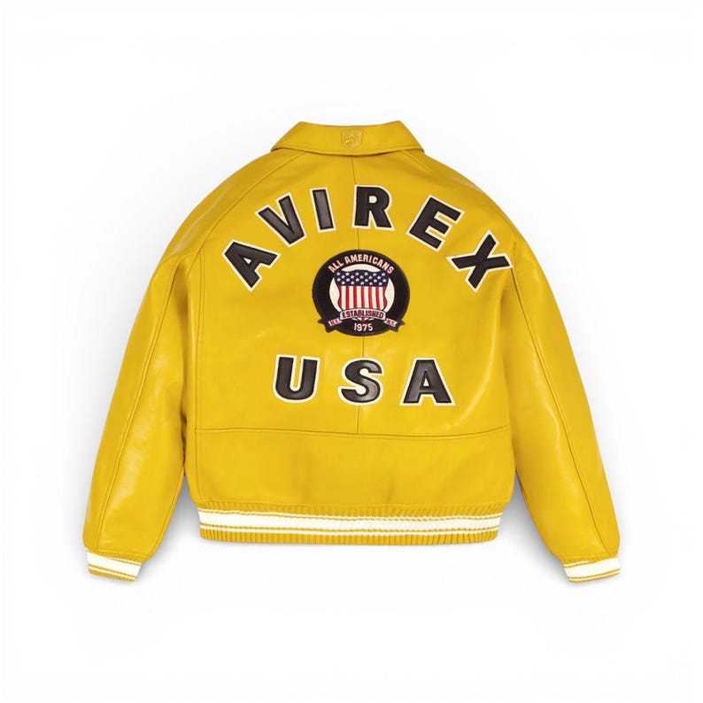 K&ouml;nnte beinhalten: Gelbe Lederjacke mit dem Wort "AVIREX" in schwarzen Buchstaben auf dem R&uuml;cken. Darunter befindet sich ein kreisf&ouml;rmiger Aufn&auml;her mit "ALL AMERICANS ESTABLISHED 1975 USA". Die Jacke hat wei&szlig;-schwarz gestreifte Manschetten.