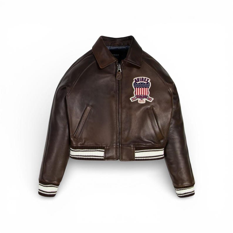 May include: A brown leather bomber jacket with a zip-up front, a collar, and a patch on the chest. The jacket has white and black striped cuffs and waistband. The patch features a crest with the text "AVIREX" and an American flag design.