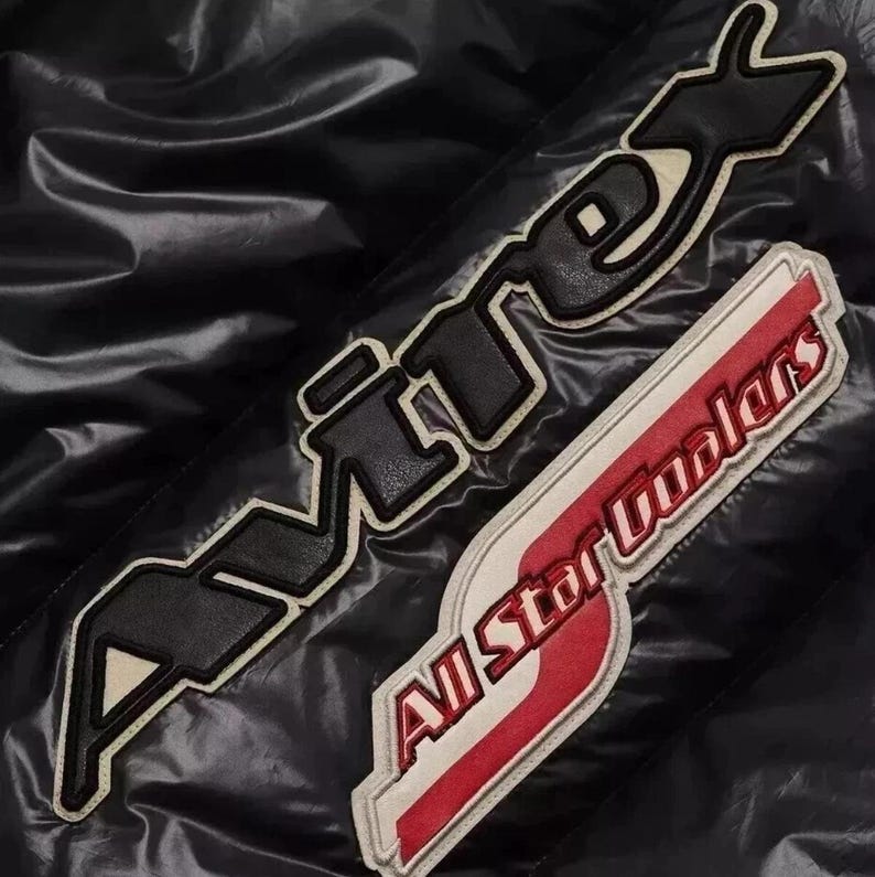 K&ouml;nnte beinhalten: Schwarze Jacke mit zwei gestickten Aufn&auml;hern. Ein Aufn&auml;her zeigt "AVIREX" in schwarzen Buchstaben mit wei&szlig;em Rand. Der zweite Aufn&auml;her zeigt "All Star Coolers" in Rot und Wei&szlig;.