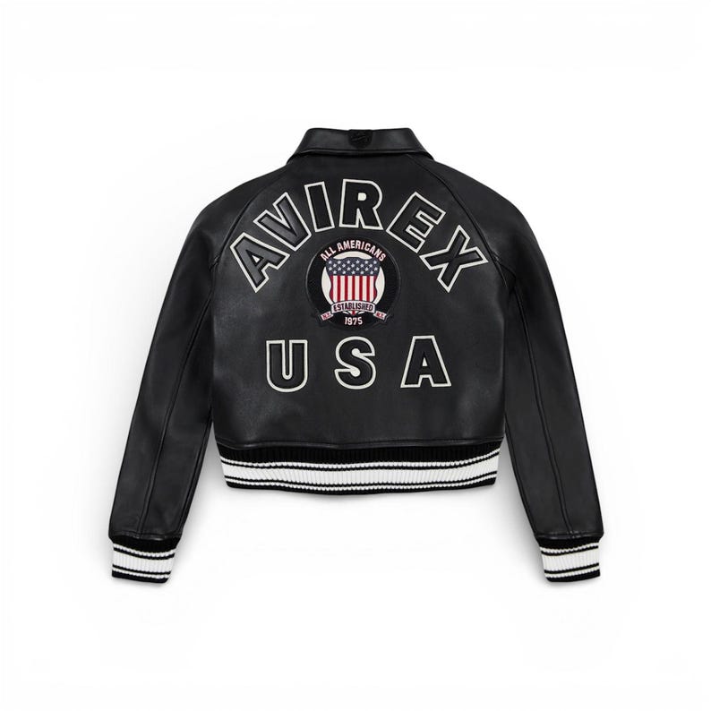 May include: Black leather jacket with white and black striped trim at the waist and cuffs. The back features the word "AVIREX" above a circular patch with the American flag and the word "USA" below.