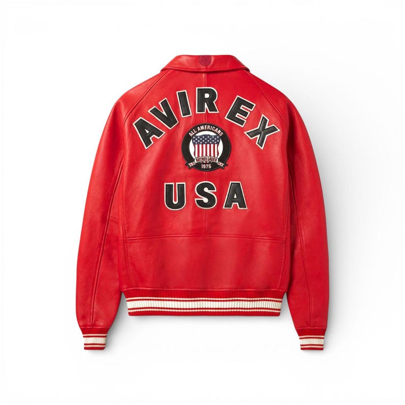 K&ouml;nnte beinhalten: Eine leuchtend rote Lederjacke mit wei&szlig;en und schwarzen Akzenten. Auf dem R&uuml;cken steht in Wei&szlig; der Schriftzug "AVIREX" und "USA", mit einem Emblem der amerikanischen Flagge. Die Jacke hat einen klassischen Bomberstil mit gerippten B&uuml;ndchen und Saum.