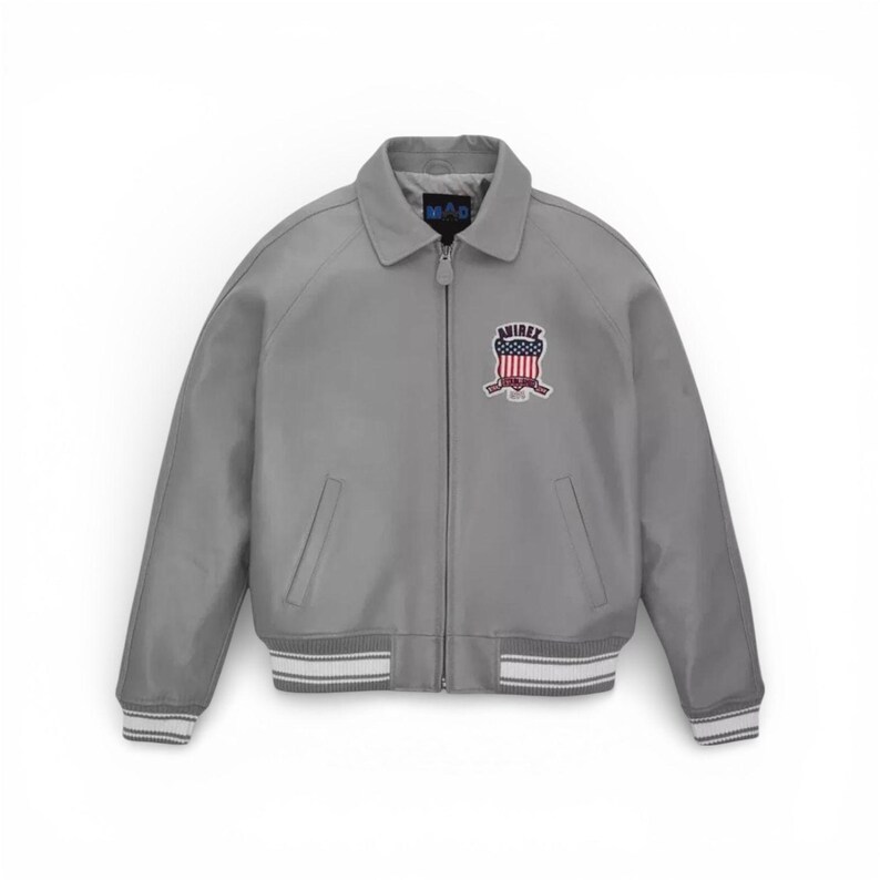 K&ouml;nnte beinhalten: Graue Bomberjacke mit Kragen und Rei&szlig;verschluss. Die Jacke hat einen Aufn&auml;her mit einem rot-wei&szlig;-blauen Design und dem Wort "AMIREX". Die Manschetten und der Saum haben wei&szlig;-graue Streifen.