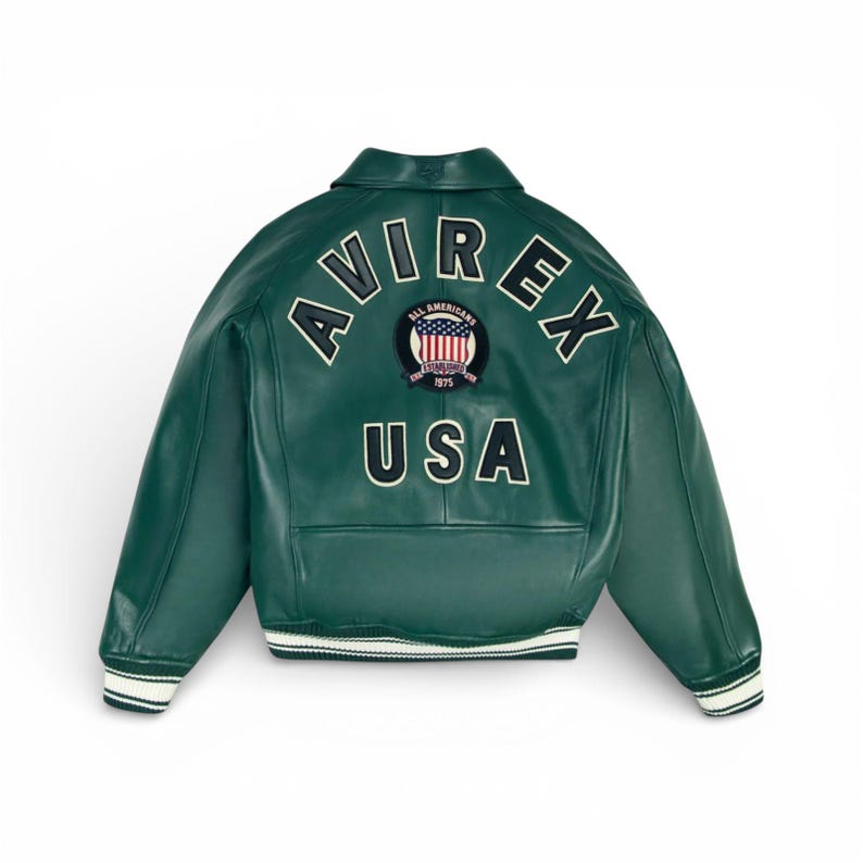 K&ouml;nnte beinhalten: Dunkelgr&uuml;ne Lederjacke mit dem Wort "AVIREX" in wei&szlig;en Buchstaben auf dem oberen R&uuml;cken. Darunter befindet sich ein kreisf&ouml;rmiger Aufn&auml;her mit der amerikanischen Flagge und dem Wort "USA". Die Jacke hat einen Kragen und wei&szlig;-schwarz gestreifte Manschetten.