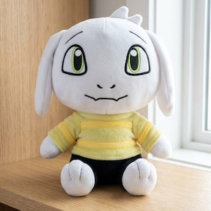 Może przedstawiać: Biała pluszowa zabawka z dużymi zielonymi oczami, długimi uszami i małym kosmykiem włosów. Nosi żółto-białą koszulę w paski i czarne szorty. Zabawka siedzi na drewnianej powierzchni.