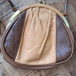 Puede incluir: Un bolso de hombro redondeado con un panel central de cuero color canela liso, flanqueado por dos paneles texturizados de color marrón oscuro con un patrón similar a la piel de serpiente. Incluye un marco de metal dorado y una correa fina.