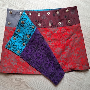 Peut inclure: Jupe patchwork avec une variété de motifs et de couleurs. La jupe présente un imprimé floral rouge, un imprimé géométrique turquoise et un imprimé cachemire violet. La ceinture est grise et bordeaux avec des œillets métalliques.