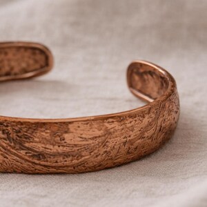 Op de afbeelding: Close-up van een koperen manchetarmband. De open armband heeft een getextureerd oppervlak en een warme, roodbruine kleur. De armband ligt op een lichtgekleurde stof en toont zijn gedetailleerde patroon.