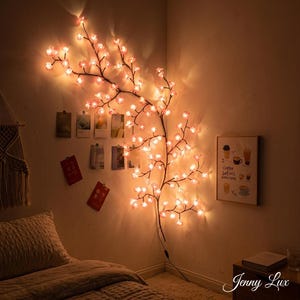 Op de afbeelding: Een decoratieve, aan de muur gemonteerde kersenbloesem-lichtslinger. De bruine takken zijn versierd met roze, verlichte bloemen. De slinger is aangesloten op een stopcontact. De muur heeft ook foto's en een print met de tekst "Coffee Solves Everything".