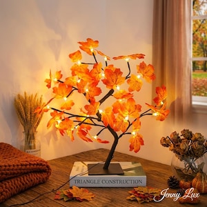 May include: A decorative tree with orange maple leaf accents and small lights. The tree sits on a square base and is placed on a wooden table. The text "TRIANGLE CONSTRUCTION" is visible on the book under the tree.