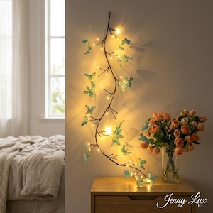 May include: A decorative wall hanging featuring a brown vine with green butterfly accents and small, warm white LED lights. The vine is mounted on a light-colored wall, with a wooden nightstand and a bouquet of roses in the foreground.