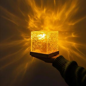 Könnte beinhalten: Eine strukturierte Würfellampe, die ein warmes gelbes Licht ausstrahlt. Die Lampe steht auf einem dunklen Holzsockel und wird in einer Hand gehalten. Das Licht erzeugt ein sternförmiges Muster auf dem Hintergrund.