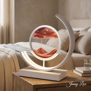 Könnte beinhalten: Eine weiße und silberne Sandkunstlampe mit kreisförmigem Glasdisplay. Das Display enthält roten Sand, der eine Landschaft bildet. Die Lampe hat einen gebogenen, beleuchteten Rahmen und einen rechteckigen Sockel. Der Text "Jenny Lux" ist sichtbar.
