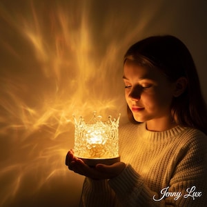 Peut inclure: Une lampe transparente en forme de couronne dégageant une lumière chaude. La lampe est tenue par une personne, projetant un motif de lumière sur le mur. La lampe a une base en bois et est étiquetée "Jenny Lux".