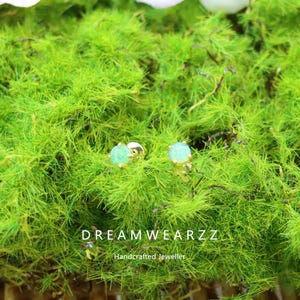 May include: A pair of gold-colored stud earrings and a ring with light green gemstones, set against a backdrop of vibrant green moss. The earrings and ring are the focal point, showcasing the jewelry's design. The brand name "DREAMWEARZZ" is visible.
