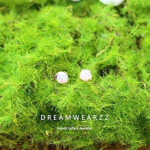 May include: A pair of round opal stud earrings with a gold setting, displayed on a bed of vibrant green moss. The earrings are the focal point, with the text "DREAMWEARZZ" and "Handcrafted Jeweller" at the bottom.
