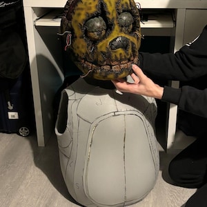 May include: A detailed, handmade Springtrap mask with a weathered, yellow and brown finish. The mask features visible wires, dark eye sockets, and a detailed mouth. A gray, unfinished costume piece is below the mask.