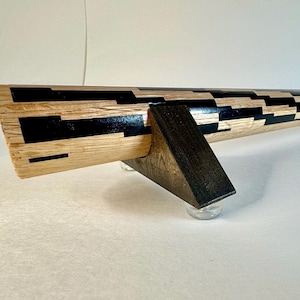 May include: A wooden decorative object with a geometric pattern of black and natural wood tones. The object is supported by two dark wooden legs and clear, rounded feet. The design is modern and artistic.