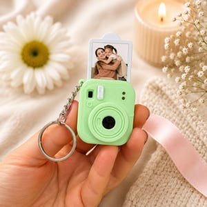 May include: A light green camera-shaped keychain with a silver chain and keyring. The camera has a small photo of two people. The keychain is held in a hand. A lit candle and flowers are in the background.