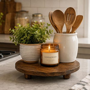 May include: A round dark wood riser with three short legs holds a white ceramic pot with a green plant, a lit amber glass candle jar labeled Sandalwood, and a white ceramic utensil crock containing four wooden spoons and a slotted turner.