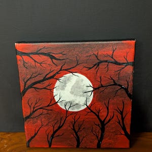 Può includere: Opera d'arte su tela quadrata con una combinazione di colori rosso e nero. L'opera raffigura una luna piena al centro, circondata da alberi a silhouette. I rami si estendono verso la luna, creando una scena drammatica e suggestiva.