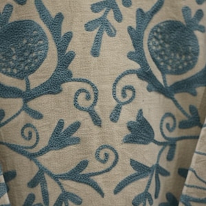 Puede incluir: Primer plano de una tela beige con patrones florales bordados en color turquesa. El dise&ntilde;o presenta flores estilizadas, hojas y zarcillos. El bordado es detallado y crea una superficie texturizada.
