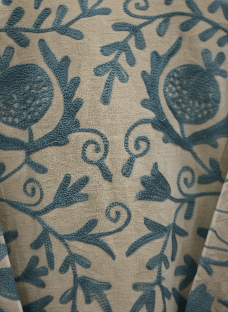 Puede incluir: Primer plano de una tela beige con patrones florales bordados en color turquesa. El dise&ntilde;o presenta flores estilizadas, hojas y zarcillos. El bordado es detallado y crea una superficie texturizada.