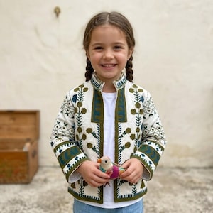 Op de afbeelding: Een witte jas met ingewikkelde borduursels in groen en blauw, met een bloemen- en geometrisch patroon. De jas heeft een opstaande kraag en wordt over een wit shirt gedragen. Het kind houdt een klein, kleurrijk vogelspeeltje vast.