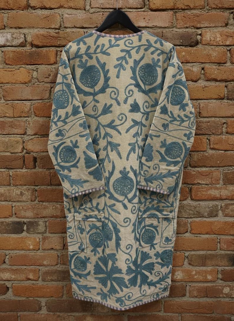 Puede incluir: Abrigo largo beige con un estampado floral turquesa y dos bolsillos. El abrigo est&aacute; colgado de una percha negra contra una pared de ladrillo. El dise&ntilde;o floral incluye motivos de granada y hojas.