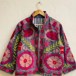 Könnte beinhalten: Eine graue Jacke mit lebendiger Blumenstickerei in Rosa, Rot und Gelb. Die Jacke hat einen Stehkragen und ein schwarz-weiß gestreiftes Futter. Das Design umfasst große kreisförmige Blumenmuster und aufwendige Blattdetails.