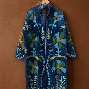 Puede incluir: Un abrigo largo de terciopelo azul oscuro con bordados florales en tonos azules, verdes y naranjas. El abrigo presenta un cuello alto y un forro a rayas. El bordado incluye flores, hojas y tallos, creando una estética bohemia.