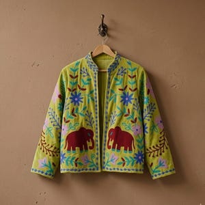Könnte beinhalten: Hellgrüne Jacke mit offenem Schnitt, verziert mit gestickten Elefanten und Blumenmustern in Rot, Blau und Lila. Die Jacke hängt an einem Holzbügel vor einer neutralen Wand.