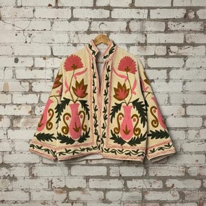 May include: Cream-coloured jacket with pink and brown floral embroidery. The jacket has a high collar and is decorated with large, stylized flowers and vase-like motifs. The embroidery is outlined in a darker pink, creating a striking contrast.
