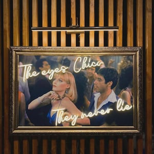 Può includere: Una stampa incorniciata con una scena di un film, con il testo al neon "The eyes Chico, They never lie". L'immagine mostra due persone in un ambiente affollato, con una cornice dorata e uno sfondo in legno.