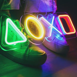 Puede incluir: Un par de zapatillas blancas con arte de luces de neón con forma de botones del mando de PlayStation: triángulo, círculo, X y cuadrado. Las luces de neón son verdes, amarillas, azules y rojas.