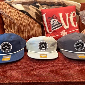 May include: Three baseball caps in shades of blue, gray, and light gray, each with a white rope detail and a circular patch featuring palm trees. The caps are displayed on a red couch with decorative pillows.