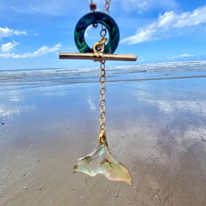 Puede incluir: Un collar dorado con cierre de palanca y un colgante con forma de cola de ballena. El colgante es iridiscente, con tonos verdes, dorados y rosas. El collar está sobre una playa y un cielo azul.